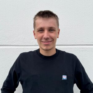 Junger Mann mit kurzem Haar lächelt, trägt schwarzes Sweatshirt vor weißer Wand.
