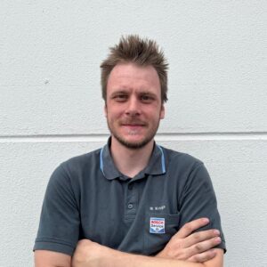 Mann lächelt, graues Bosch-Poloshirt, kurzer Bart, stacheliges Haar, vor weißer Wand.